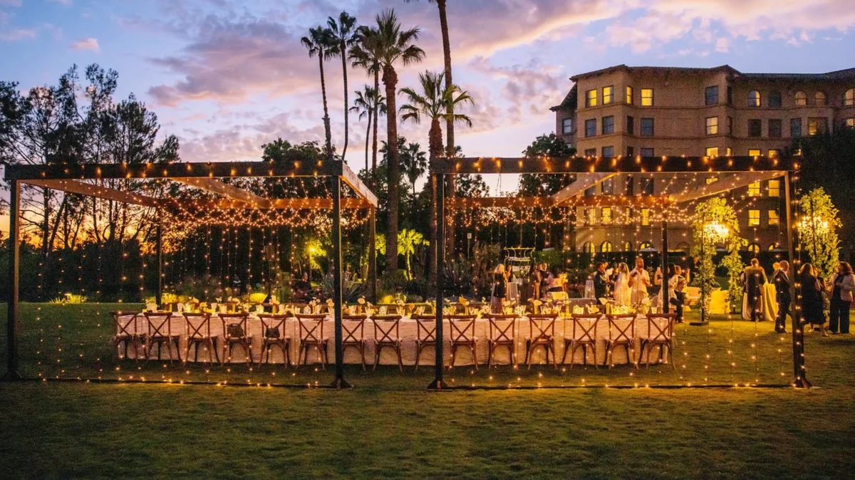 Outdoor corporate event at dusk with long banquet table under string-light canopy