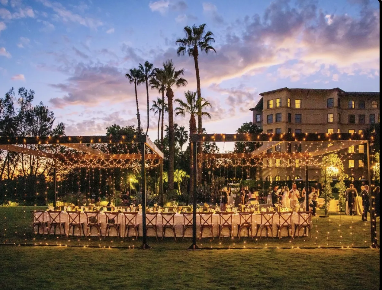 Long banquet table at sunset framed by palm trees and string-light pergola on the Langham lawn