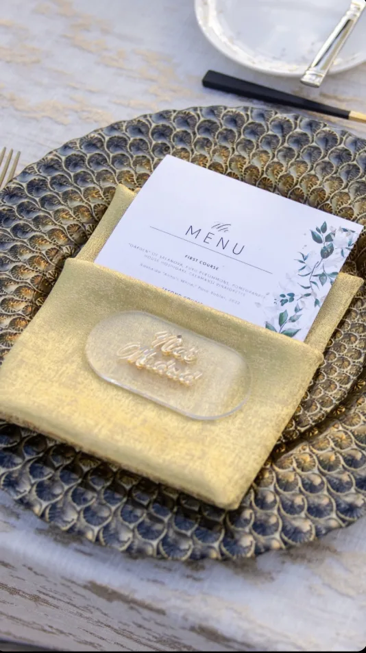 Place setting with gold napkin, printed menu card, and custom acrylic name place card on a beaded charger